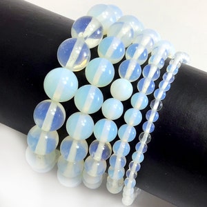 May include: A set of five bracelets made with white opal beads. The bracelets are stacked on a black surface. The beads are round and have a smooth, polished finish.
