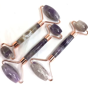 May include: Three amethyst face rollers with rose gold metal frames. The rollers are made of natural amethyst and have a purple and white color.