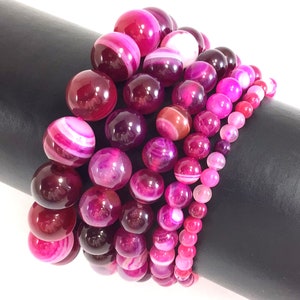 May include: A set of six pink agate gemstone bracelets with varying patterns and shades of pink. The bracelets are stacked on a black surface.