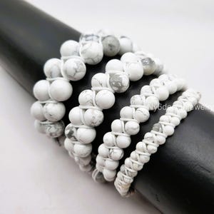 May include: Five white howlite beaded bracelets stacked on a black surface. The bracelets are made with white howlite beads and white string.
