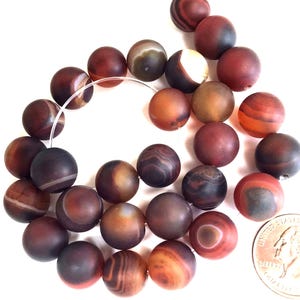 May include: A collection of matte, round beads in shades of brown, red, and orange, with unique banded patterns. The beads are strung on a clear cord, and a U.S. quarter is included for size reference. These beads are suitable for jewelry making.
