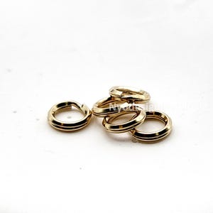 May include: A collection of gold-colored jump rings, each with a rounded, flat profile and a single, thin groove. The rings are scattered on a white surface, with some overlapping. The jewelry findings are for crafting.