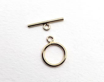 May include: A gold-colored toggle clasp set, consisting of a circular ring and a bar with a loop. The jewelry findings are displayed against a white background. The clasp is likely used for necklaces or bracelets.