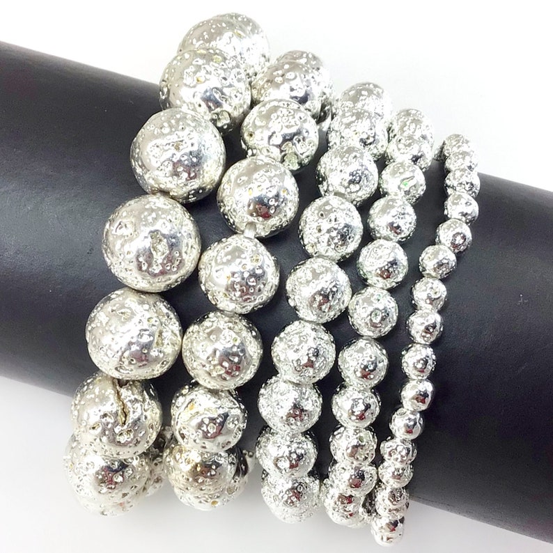 May include: A collection of silver-colored beaded bracelets. The beads have a textured, pitted surface, and vary in size. The bracelets are arranged on a black cylindrical form, showcasing the jewelry's design and texture. The beads reflect light, creating a shiny appearance.