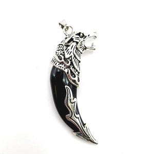 May include: A silver-colored wolf head pendant with a black, curved tooth design. The wolf head features detailed fur and an open mouth. The pendant is designed to be worn as a necklace.