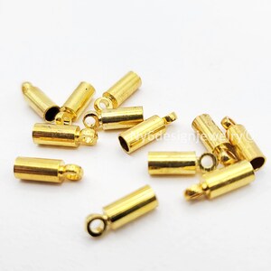 May include: A collection of gold-colored jewelry findings, including cylindrical crimp end caps with small loops and open-ended tubes. These components are designed for jewelry making and crafting projects. The findings are scattered on a white surface.