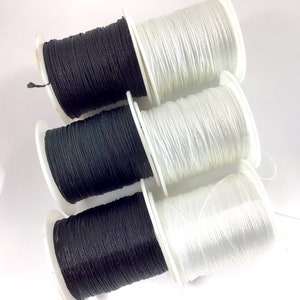 May include: Six spools of thread, three black and three white, are wound on white plastic bobbins. The spools are arranged in two stacks of three, with the black spools on the bottom and the white spools on top.