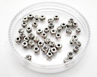 May include: A collection of small, silver-colored, geometric beads with a hexagonal shape and decorative patterns. The beads are contained within a clear, round plastic container, and are scattered throughout. The beads are likely used for jewelry making or crafting.