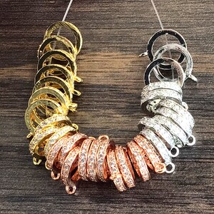 K&ouml;nnte beinhalten: Nahaufnahme von drei S&auml;tzen Schmuckverschl&uuml;ssen. Die Verschl&uuml;sse sind aus Metall und haben eine runde Form. Die Verschl&uuml;sse sind mit kleinen, klaren Steinen verziert. Die Verschl&uuml;sse sind in Gold, Ros&eacute;gold und Silber.