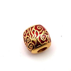 May include: A gold-colored bead with a red, swirling pattern. The bead has a cylindrical shape with a hole running through the center. The red design is set against a gold background.