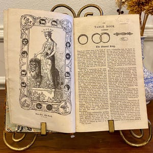 May include: An open antique book, held by a decorative gold stand, displays illustrations and text. The book's pages feature detailed drawings and the title "The Table Book." A vase with dried flowers is visible in the background.