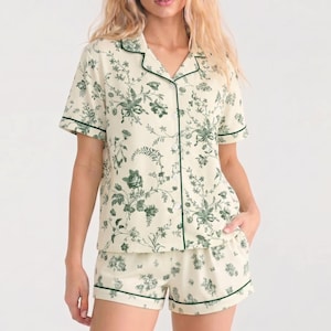Könnte beinhalten: Ein zweiteiliger Schlafanzug bestehend aus einem kurzärmeligen Hemd mit Knopfleiste und passender Shorts. Das Set ist cremefarben mit einem dunkelgrünen floralen Toile-Muster und dunkelgrünen Paspeln an Kragen, Knopfleiste und Säumen.