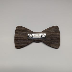 May include: A wooden bow tie with a dark brown wood grain pattern. A silver metal plate in the center reads "CAUTION MAGNETS" and has two small triangles on each side. The bow tie is a classic accessory.