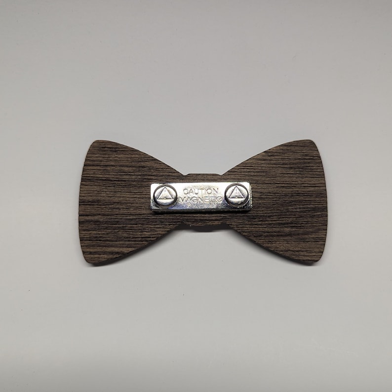 May include: A wooden bow tie with a dark brown wood grain pattern. A silver metal plate in the center reads "CAUTION MAGNETS" and has two small triangles on each side. The bow tie is a classic accessory.