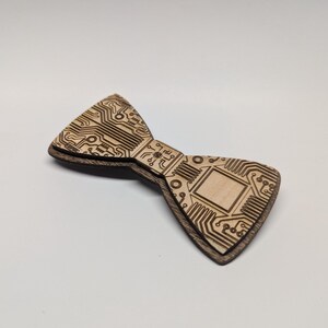 May include: A wooden bow tie with a circuit board design. The bow tie is made of two layers of wood, with the top layer featuring etched lines and shapes resembling a circuit board. The bottom layer is a darker brown.