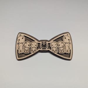 May include: Wooden bow tie featuring a laser-cut design. The bow tie has a dark brown border and a lighter wood interior with a design of several small figures and a central mask.