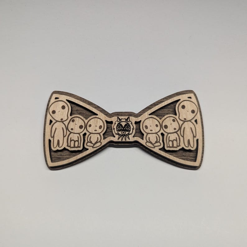 May include: Wooden bow tie featuring a laser-cut design. The bow tie has a dark brown border and a lighter wood interior with a design of several small figures and a central mask.