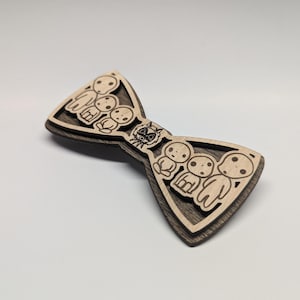 May include: Wooden bow tie featuring a layered design with a light wood top layer and a dark wood base. The bow tie has a laser-cut design of several small figures and a circular character in the center.