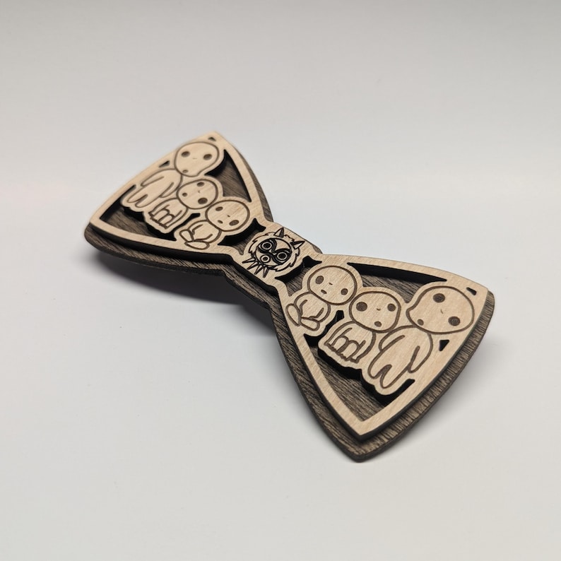 May include: Wooden bow tie featuring a layered design with a light wood top layer and a dark wood base. The bow tie has a laser-cut design of several small figures and a circular character in the center.