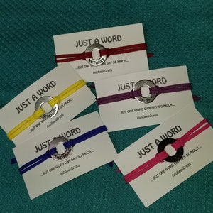 May include: A collection of handmade bracelets with a metal washer centerpiece and colorful cords. Each bracelet is displayed on a card with the text "JUST A WORD" and a motivational word like "STRENGTH" or "PERSEVERANCE".