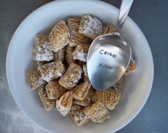 May include: A white bowl filled with frosted cereal and a silver spoon with the words "Cereal Killer" engraved on it.