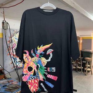 Op de afbeelding: Zwart t-shirt met een kleurrijk, abstract schedelontwerp. De afbeelding bevat een mix van heldere kleuren, waaronder rood, blauw, geel en groen. Het shirt hangt aan een witte hanger.
