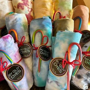 May include: A woven basket filled with rolled tie-dye towels in various colors, including blue, pink, and yellow. Each towel is tied with a red ribbon and a candy cane. The towels have tags that read "RY GUY DYES".