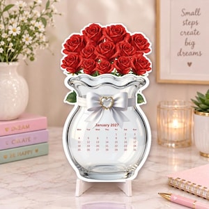May include: A decorative desk calendar shaped like a clear glass vase filled with red roses. A white ribbon with a heart-shaped charm is tied around the neck of the vase. The calendar displays the month of January 2027 with dates in red text.