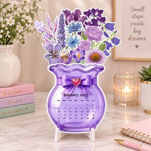 May include: A purple vase-shaped desk calendar for January 2027, featuring a vibrant bouquet of purple, lavender, and blue flowers. The vase is decorated with a purple ribbon and a small heart-shaped charm, supported by a white stand.