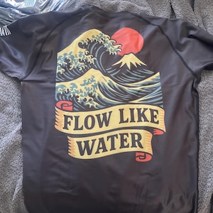 May include: Black short-sleeved shirt featuring a Japanese wave design with a red sun and a mountain. The shirt has the text "FLOW LIKE WATER" on a banner. The design is in shades of blue, yellow, and red.