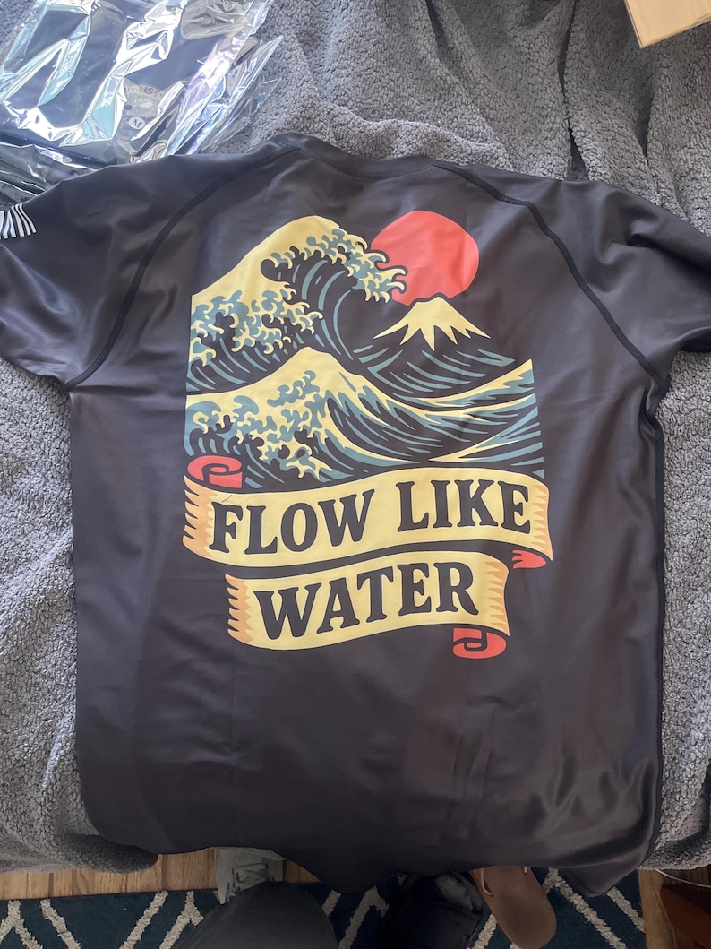 May include: Black short-sleeved shirt featuring a Japanese wave design with a red sun and a mountain. The shirt has the text "FLOW LIKE WATER" on a banner. The design is in shades of blue, yellow, and red.