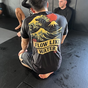 May include: Black t-shirt with a graphic of a wave, mountain, and red sun design. The text "FLOW LIKE WATER" is printed in yellow. The shirt is worn by a person kneeling on a black mat.