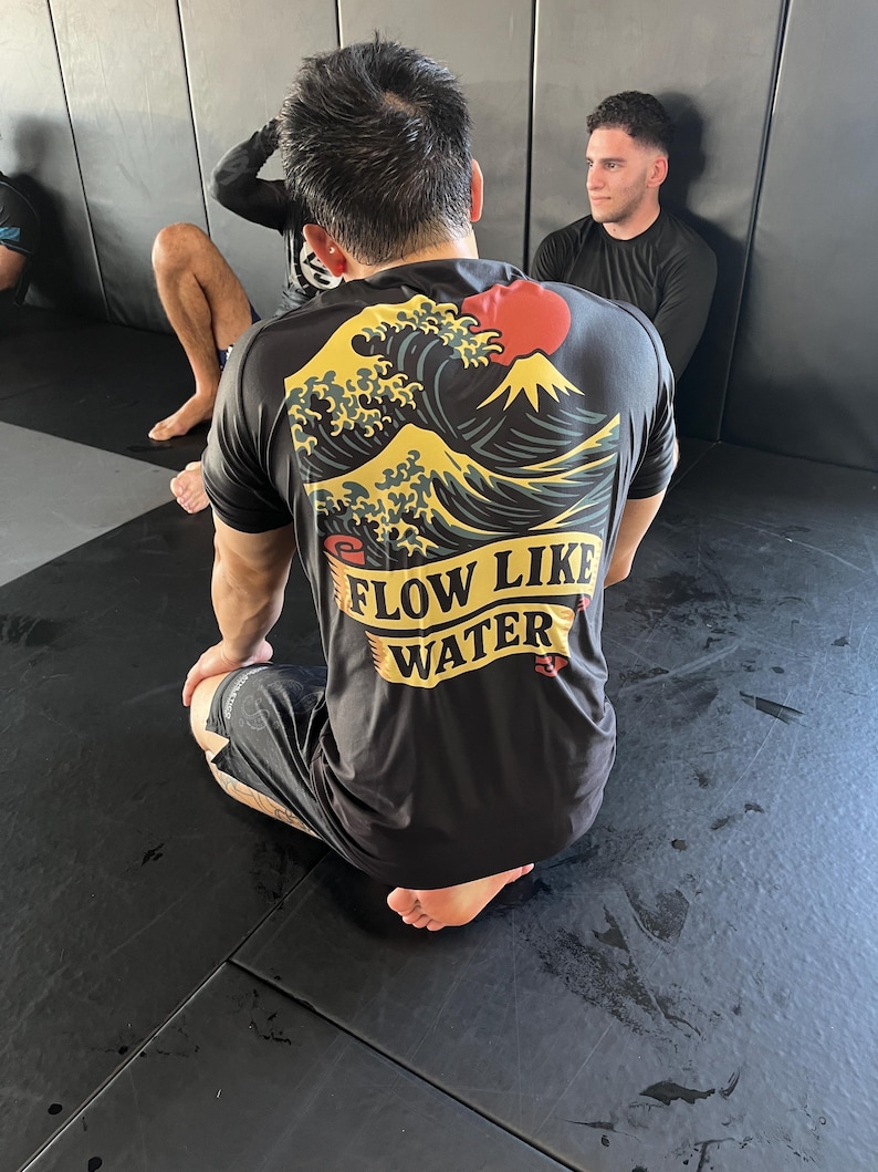 May include: Black t-shirt with a graphic of a wave, mountain, and red sun design. The text "FLOW LIKE WATER" is printed in yellow. The shirt is worn by a person kneeling on a black mat.