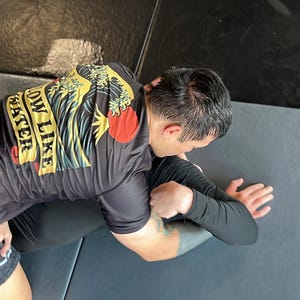 May include: A black t-shirt with the text "FLOW LIKE WATER" in yellow and a red sun graphic. The shirt is worn by a person in a grappling position, with black shorts and a black long-sleeve shirt. The scene takes place on a gray mat.