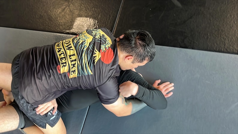 May include: A black t-shirt with the text "FLOW LIKE WATER" in yellow and a red sun graphic. The shirt is worn by a person in a grappling position, with black shorts and a black long-sleeve shirt. The scene takes place on a gray mat.