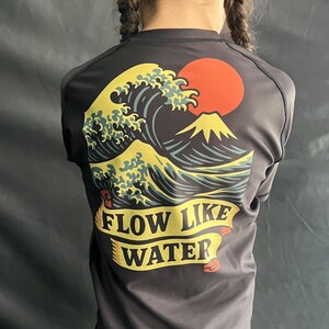 May include: Dark gray athletic shirt featuring a graphic of a large ocean wave, a red sun, and a mountain. The text "FLOW LIKE WATER" is displayed on a yellow banner. The shirt has a crew neck and short sleeves.