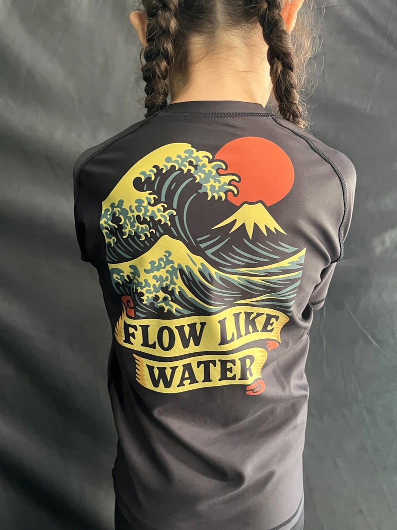 May include: Dark gray athletic shirt featuring a graphic of a large ocean wave, a red sun, and a mountain. The text "FLOW LIKE WATER" is displayed on a yellow banner. The shirt has a crew neck and short sleeves.