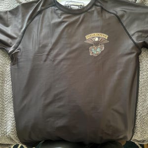 May include: A dark gray short-sleeve shirt with black trim and a white collar. The shirt features a "FIDELIS DEFENSE" emblem with an eagle, globe, and anchor design on the chest. The shirt is made of a smooth, possibly athletic material.