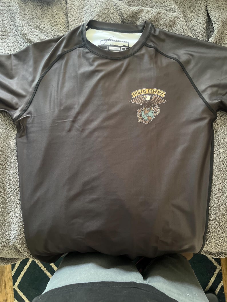 May include: A dark gray short-sleeve shirt with black trim and a white collar. The shirt features a "FIDELIS DEFENSE" emblem with an eagle, globe, and anchor design on the chest. The shirt is made of a smooth, possibly athletic material.