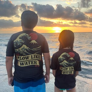 May include: Two black t-shirts with a Japanese wave and mountain design and the text "FLOW LIKE WATER". The shirts are worn by two people standing on a beach at sunset. One person wears blue shorts.