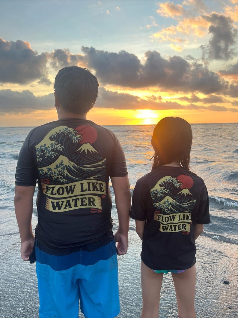 May include: Two black t-shirts with a Japanese wave and mountain design and the text "FLOW LIKE WATER". The shirts are worn by two people standing on a beach at sunset. One person wears blue shorts.