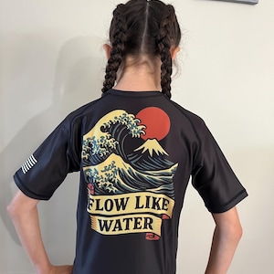 May include: A dark gray short-sleeved shirt with a graphic of a wave, mountain, and red sun. The text "FLOW LIKE WATER" is displayed on a banner. The shirt has black sleeves and a white stripe design on the left sleeve.