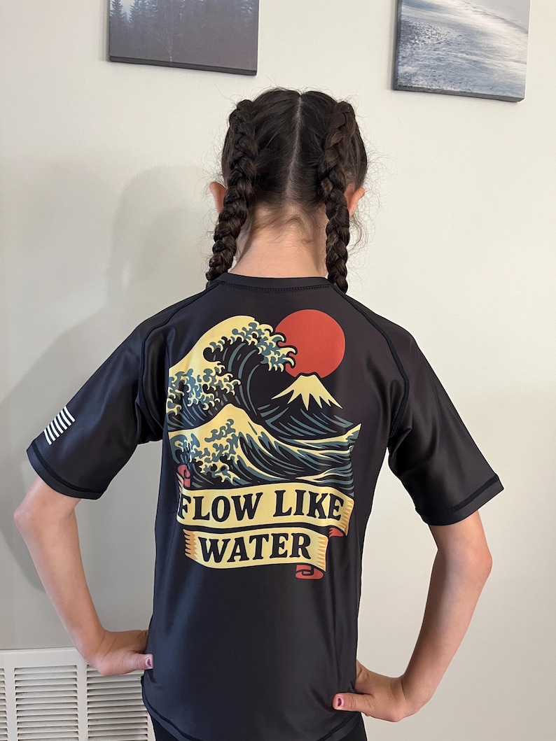 May include: A dark gray short-sleeved shirt with a graphic of a wave, mountain, and red sun. The text "FLOW LIKE WATER" is displayed on a banner. The shirt has black sleeves and a white stripe design on the left sleeve.