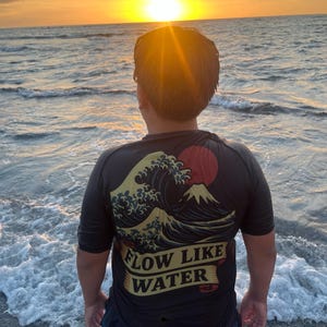 May include: A person wearing a black t-shirt with a Japanese wave and mountain design, and the text "FLOW LIKE WATER". The person is standing on a beach, facing the ocean at sunset. The sky is a mix of orange and yellow.