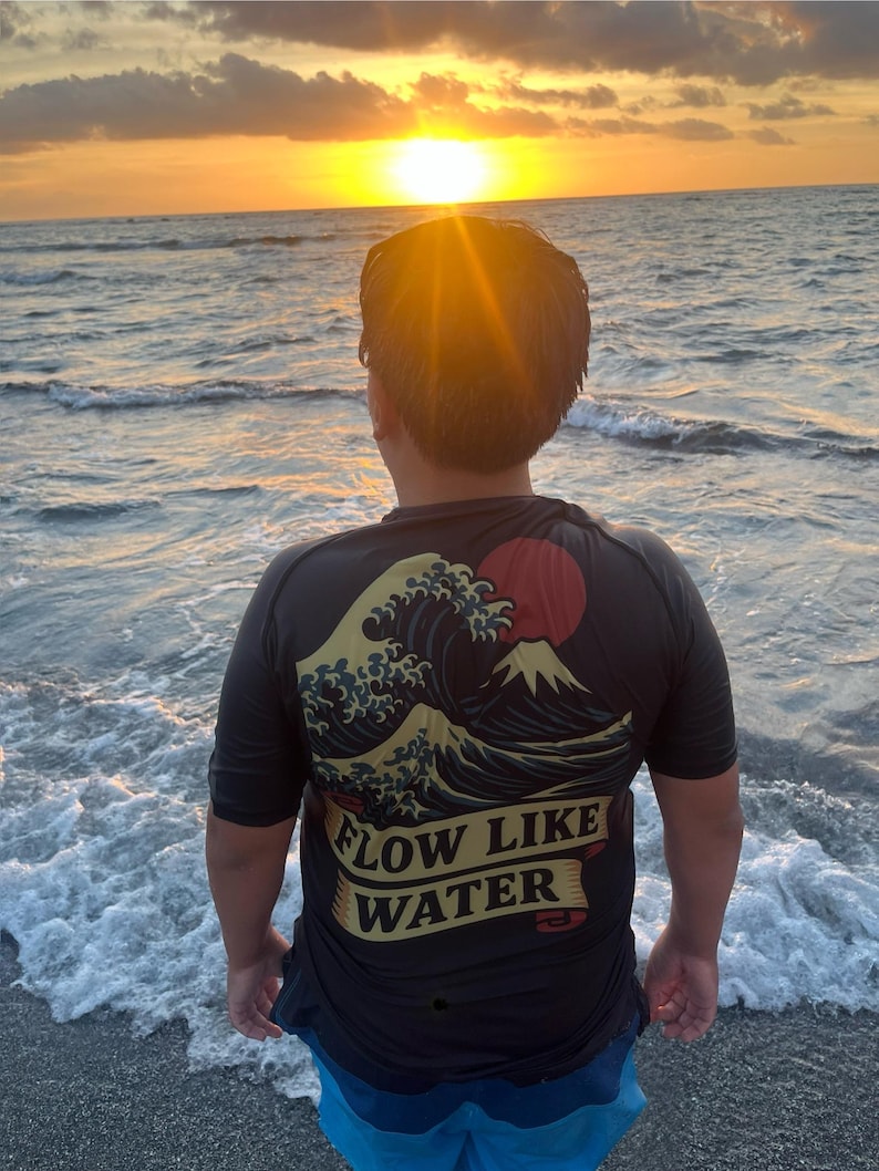 May include: A person wearing a black t-shirt with a Japanese wave and mountain design, and the text "FLOW LIKE WATER". The person is standing on a beach, facing the ocean at sunset. The sky is a mix of orange and yellow.