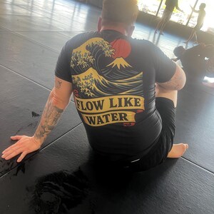 May include: Black athletic shirt with a Japanese wave and mountain graphic, and the text "FLOW LIKE WATER" in a banner. The shirt is worn by a person sitting on a black mat in a gym setting.