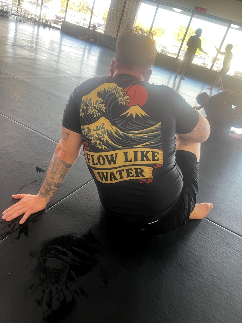 May include: Black athletic shirt with a Japanese wave and mountain graphic, and the text "FLOW LIKE WATER" in a banner. The shirt is worn by a person sitting on a black mat in a gym setting.