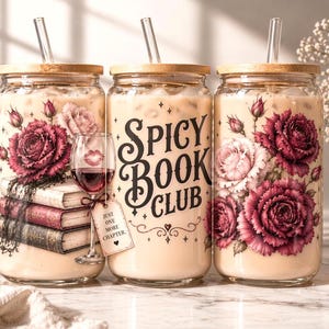 May include: Three glass tumblers with wooden lids and glass straws, filled with a creamy beverage. Each tumbler features a floral design with burgundy and pink roses. The middle tumbler has the text "Spicy Book Club". A wine glass with red wine and a tag that says "Just one more chapter" is in front.