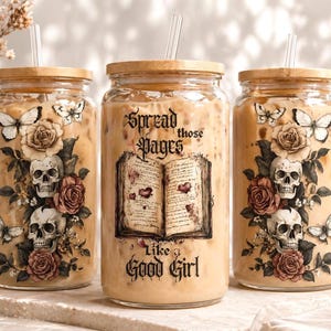 May include: Three glass mason jars with wooden lids, filled with a creamy beverage and straws. The jars are decorated with skulls, roses, butterflies, and the text "Spread those Pages Like a Good Girl."