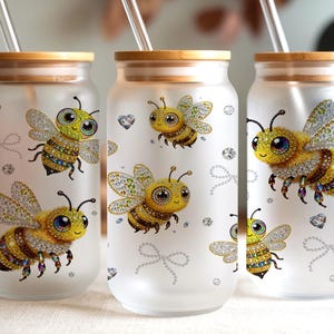 May include: Three frosted glass tumblers with bamboo lids and clear straws. Each tumbler features a detailed illustration of a cartoon bee with yellow, black, and white accents, along with decorative rhinestone embellishments. The tumblers are designed for cold beverages.
